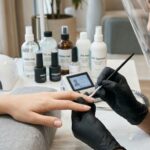 A focused professional nail technician wears black gloves and a face shield while performing a detailed manicure with a fine brush. Bottles labeled HEMA-Free, TPO-Free, and SDS-Approved are on the modern workstation alongside a UV lamp.